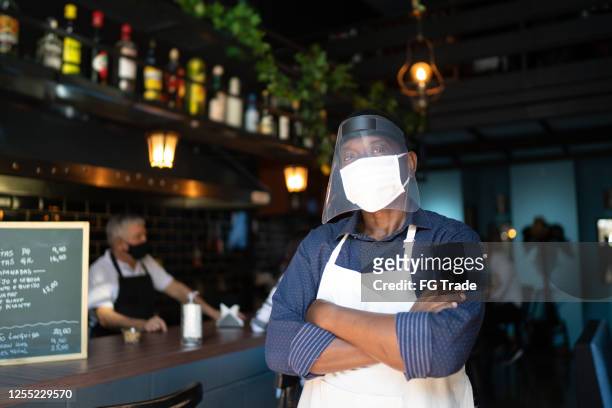 porträt eines kellners mit gekreuzten armen und schutzischer gesichtsmaske im restaurant - essenzielle berufe und dienstleistungen stock-fotos und bilder