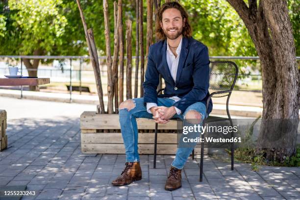 smiling handsome young male professional sitting with hands clasped at cafe - ripped jeans stock pictures, royalty-free photos & images