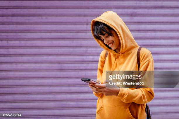 portrait of young woman with yellow hoodie checking smartphone in front of purple background - young adult imagens e fotografias de stock