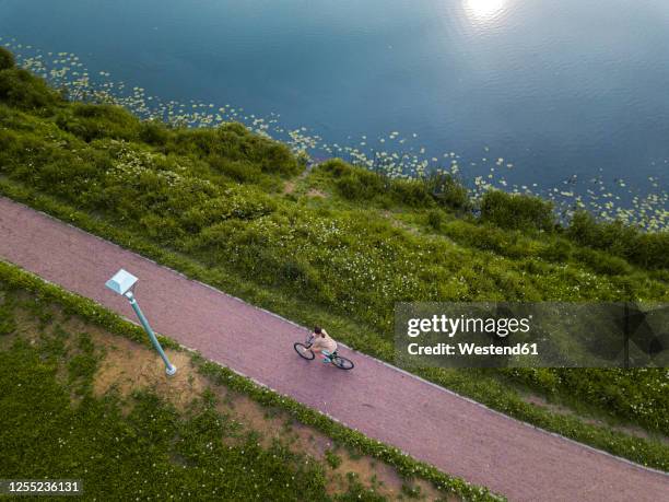 russia, leningrad oblast, tikhvin, aerial view of female cyclist riding along lakeshore footpath in park - lakeshore stockfoto's en -beelden