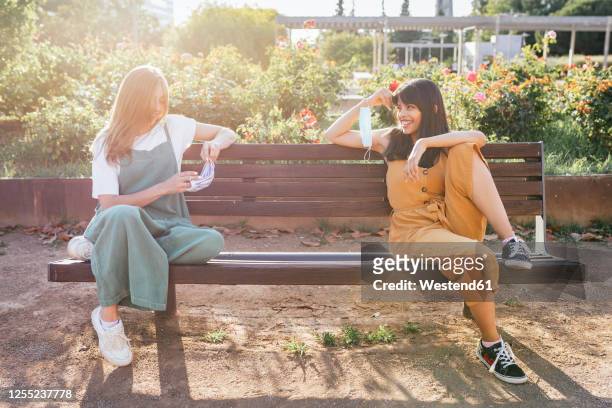 two friends with protective masks sitting on bench during corona crisis keeping distance - social distancing stock pictures, royalty-free photos & images