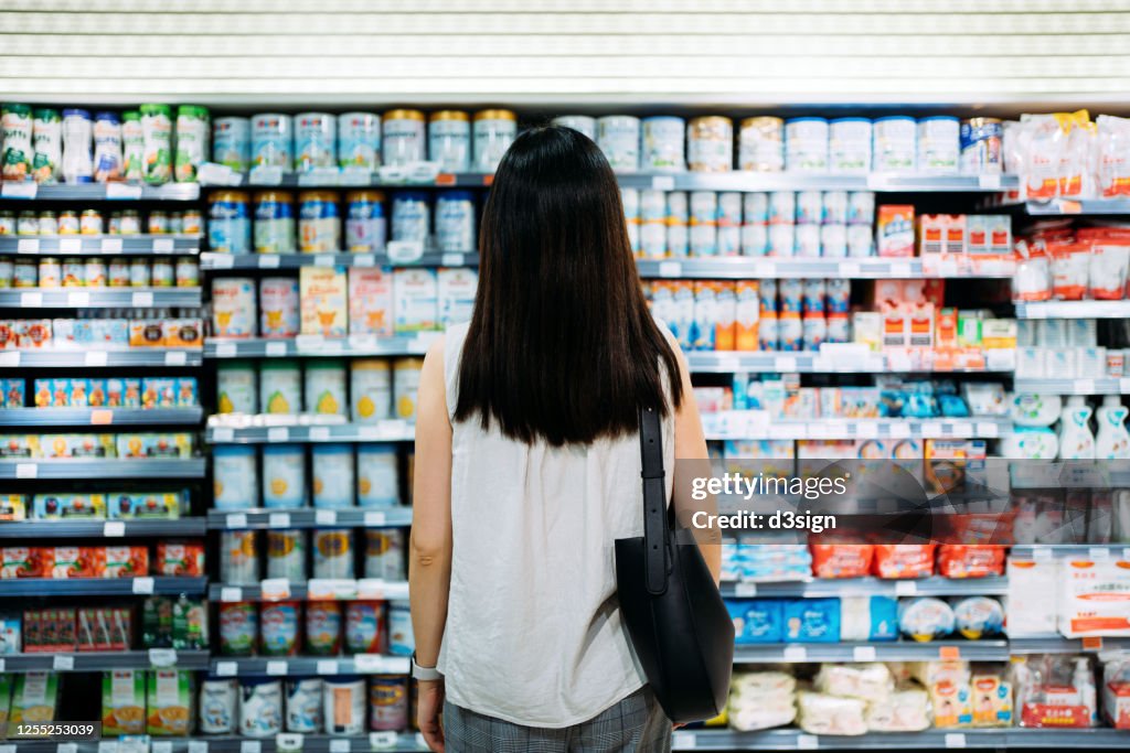 Rear view of young Asian mother groceries shopping for baby products in a supermarket. She is standing in front of the baby product aisle and have no idea which product to choose from