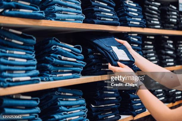 cropped shot of woman's hand selecting a pair of trousers from the display shelf while shopping in a clothing store in the city - kleidungsstück stock-fotos und bilder