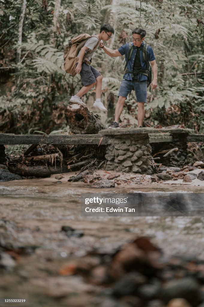 An asian chinese father hiking and exploring forest helping his son crossing the bridge on the river