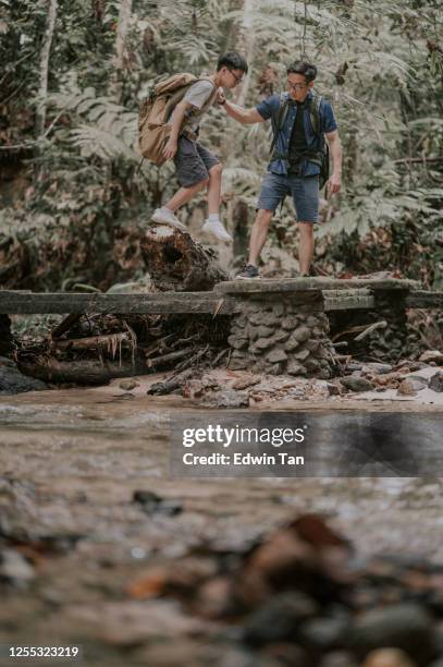 an asian chinese father hiking and exploring forest helping his son crossing the bridge on the river - helping to cross the bridge stock pictures, royalty-free photos & images