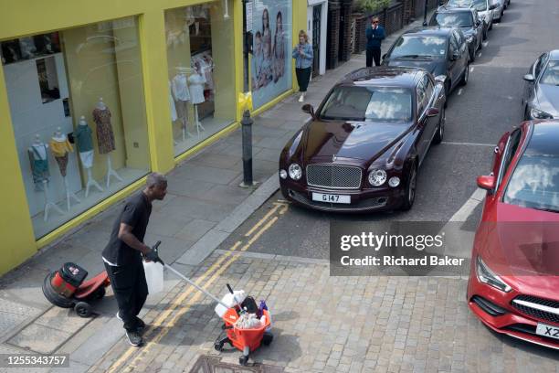 Contact cleaner with work equipment crosses the road in front of luxury Bentley and Mercedes cars on a side street off the King's Road in Chelsea, on...
