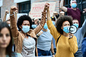 Black female activists holding hands while protesting with crowd of people during coronavirus pandemic.