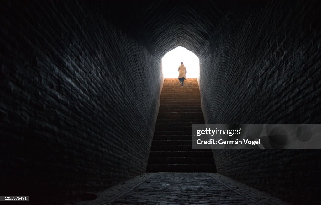 Woman walks confidently up stairs coming out from dark tunnel into bright light, Yazd, Iran