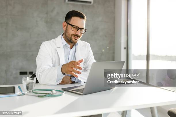 especialista médico sorridente tendo uma chamada de vídeo sobre laptop no escritório. - webconferência - fotografias e filmes do acervo