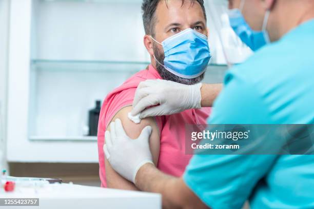 patient avec le masque protecteur regardant le docteur désinfectant son bras pour la vaccination - coton tige photos et images de collection