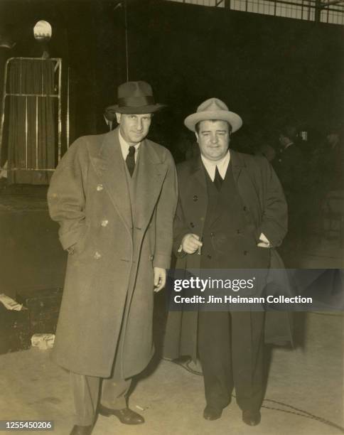 American comedy duo Bud Abbott and Lou Costello, both wearing suits and long coats, pose inside a high-ceiling building during World War II, circa...