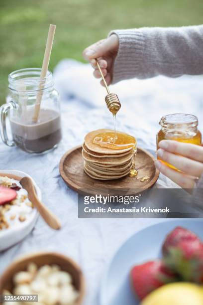 summer healthy vegetarian picnic in park.
womans hand pouring organic honey on delicious stack of pancakes.
fruits, cacao, honey pancakes and organic granola on linen tablecloth decorated with pillows and wicker basket. - crepe textile fotografías e imágenes de stock
