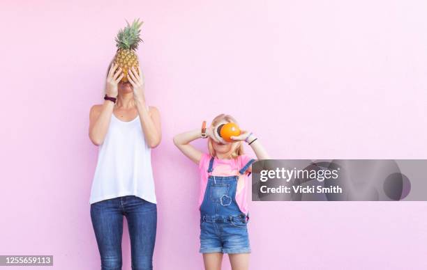 young adult mother and daughter holding fruit in front of their face against a pink wall - family portrait humor fotografías e imágenes de stock