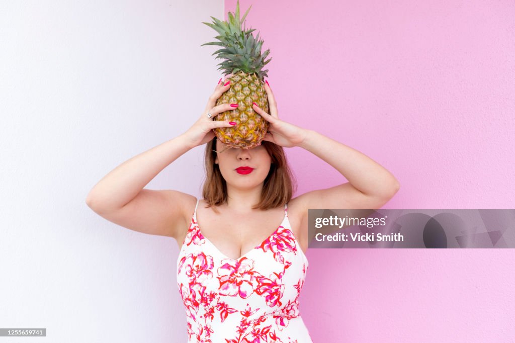 Young Woman in a red and white patterned dress, red lipstick , standing against a pink and white wall holding a pineapple in front of her face