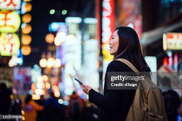 young asian female traveller with backpack using smartphone while exploring and strolling in busy local city street at night in osaka, japan - tokyo fashion stock pictures, royalty-free photos & images