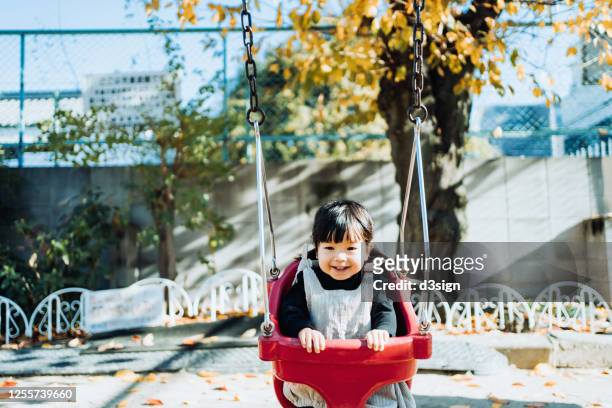 adorable little asian girl playing on swing and smiling joyfully in an outdoor playground on a lovely sunny day - área de juego fotografías e imágenes de stock