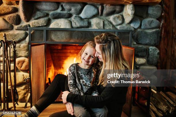 smiling mother and daughter sitting together in front of fireplace in winter cabin - log cabin fire stock pictures, royalty-free photos & images