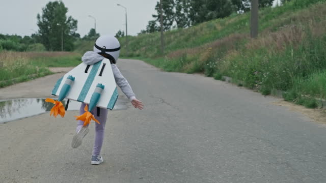 Little girl wearing white helmet, jetpack and running on rural road like aviator