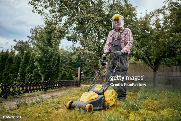 actieve hogere mens in overalls en met bescherming die het gras in zijn binnenplaats met duwmaaier maait - handgrasmaaier stockfoto's en -beelden