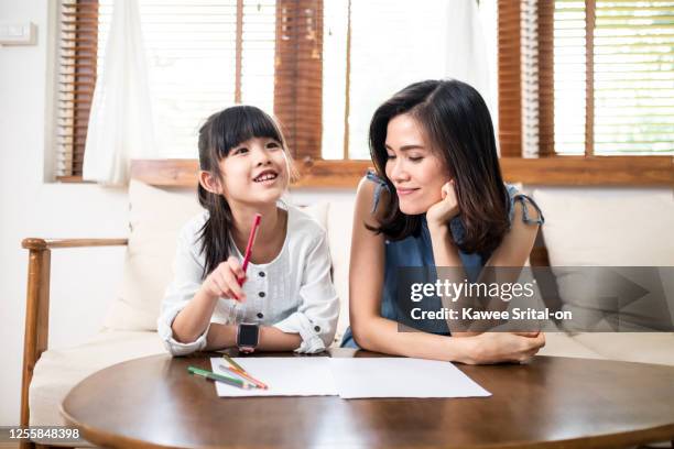 homeschool asian young little girl drawing and painting on paper on table. mother sit on sofa and give her encouragement, teach and play with her in living room. daughter smile and happy to study at home together with mom. - smile woman child stock-fotos und bilder
