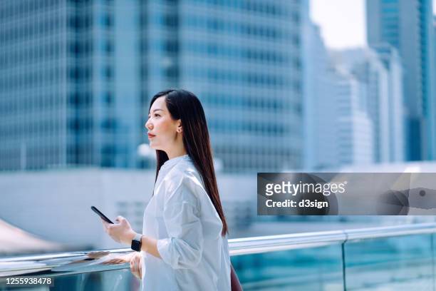 confidence young asian businesswoman holding smartphone standing on the urban balcony of office building looking over the city and planning about the future - city future ストックフォトと画像