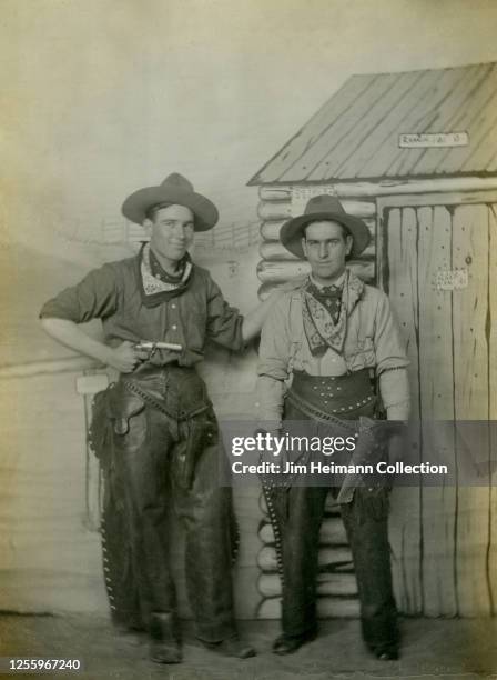 Two men pose in cowboy outfits in front of a painted backdrop depicting the Old West, 1926.