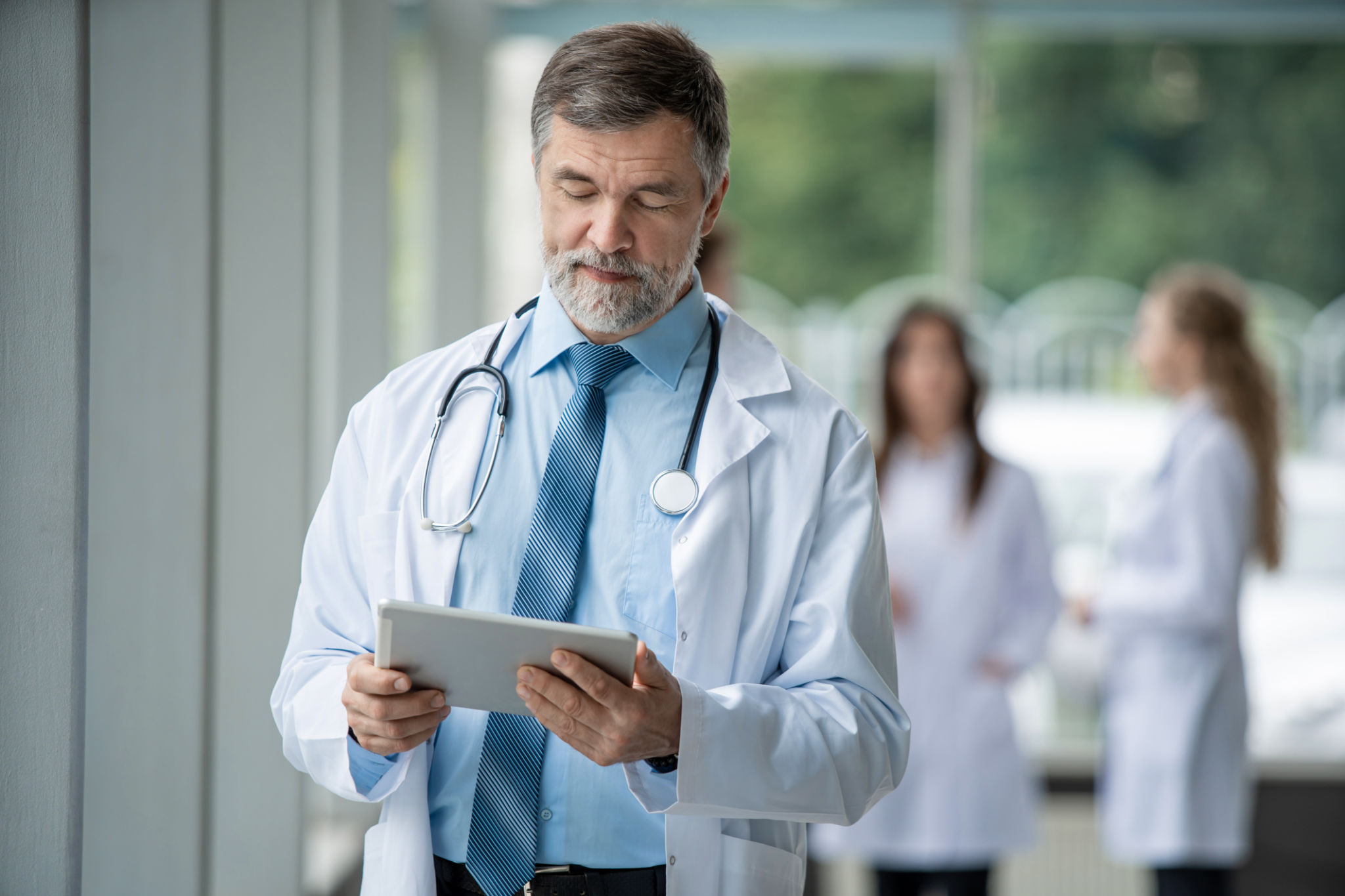 Confident smiling doctor standing with a tablet and looking at camera in a hospital. Medical team standing behind doctor Confident smiling doctor standing with a tablet and looking at camera in a hospital. Medical team standing behind doctor