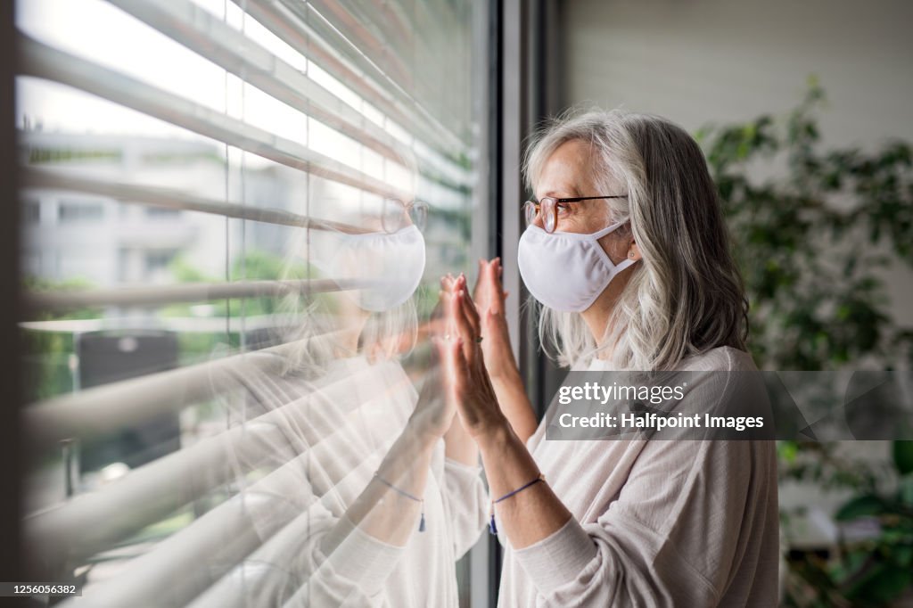 Portrait of senior woman with face mask standing indoors by window at home, looking out.