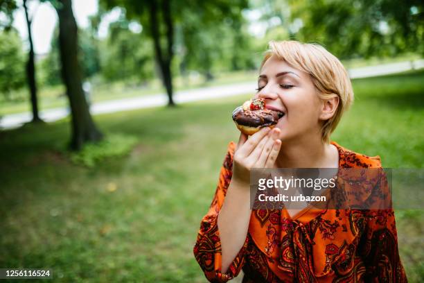 woman eating donut in park - blond women happy eating stock pictures, royalty-free photos & images