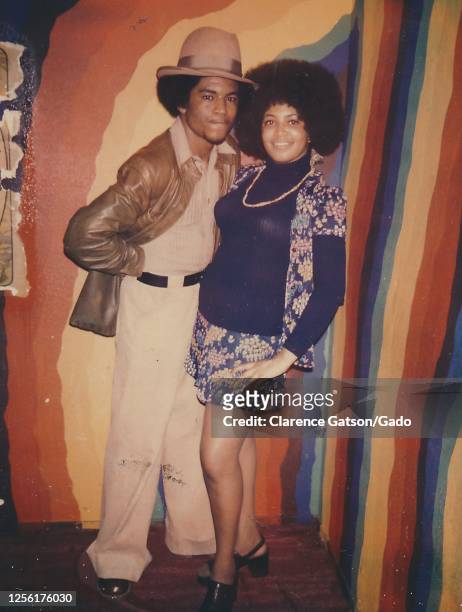 African-American couple, with the man wearing a brimmed hat and the woman wearing an afro haircut, standing arm in arm in front of a rainbow...