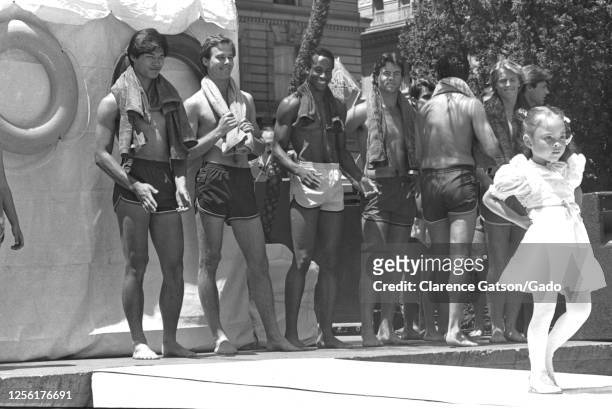 Black and white photograph a young girl wearing a white dress, modeling on a catwalk, a group of young men in the background, wearing shorts and...