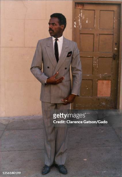 Portrait of an African-American man standing in front of a wooden doorway, wearing a tailored gray suit, with one hand on his chest, looking to the...