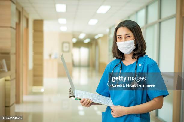 shot of asian female nurse holding a medical record in an hospital corridor. her wearing protective face mask and hospital gown for prevention infection virus. - hospital gown fotografías e imágenes de stock