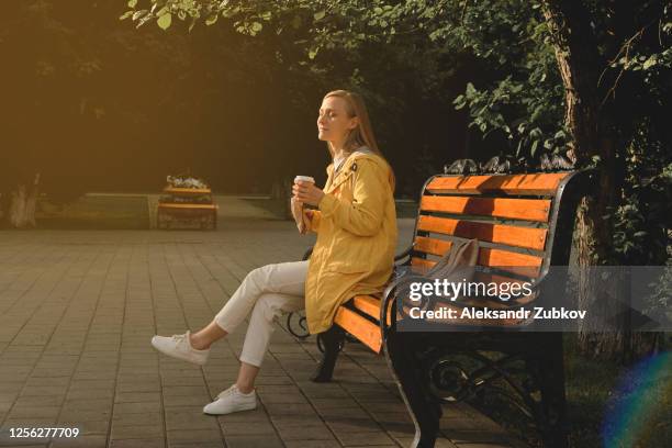 a young woman or girl holding a cup of coffee, drinking it, eating a delicious shawarma doner kebab, sitting on a bench in a public park. the concept of a fast food lunch outdoors. - adult girl single park photos et images de collection