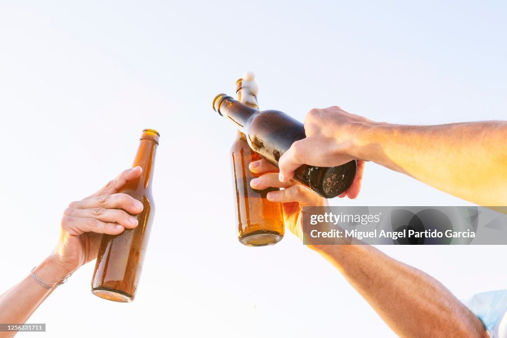 Three people doing celebratory toast with beer bottles on beach at sunset, Oliva, Valencia, Spain. - stock photo