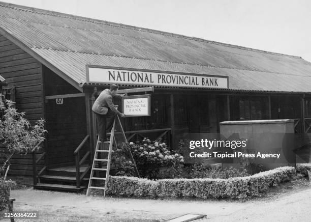 Workman hanging the sign outside the National Provincial Bank in the former military convalescent camp which will be home to 1,500 Olympic visitors...