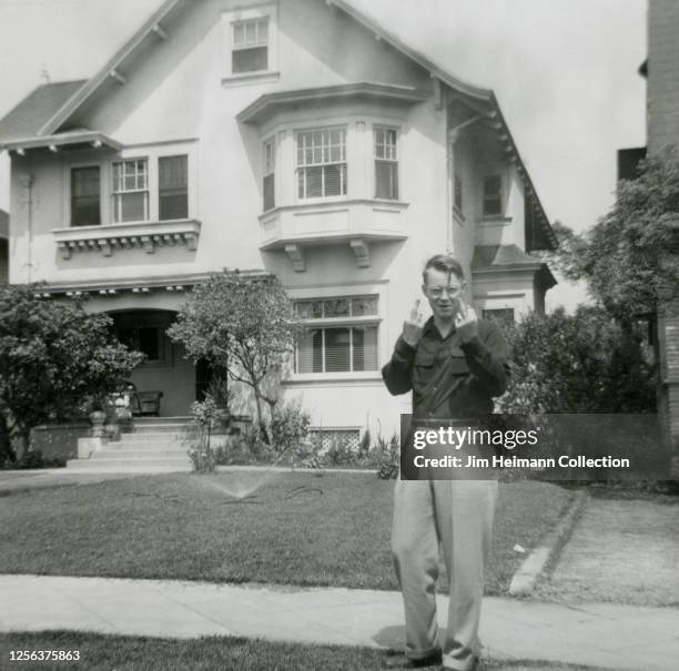 Clean-cut young man standing in front a two-story house in Los Angeles sticks up his two middle fingers while smirking at the camera, circa 1948.