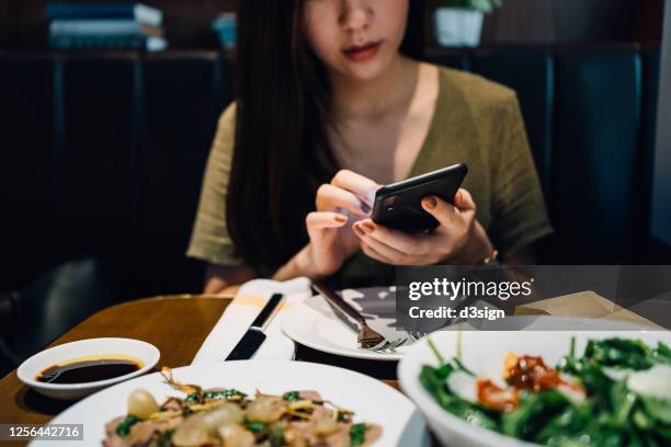 concentrated young asian woman using smartphone while having meal in a restaurant - paying for dinner imagens e fotografias de stock