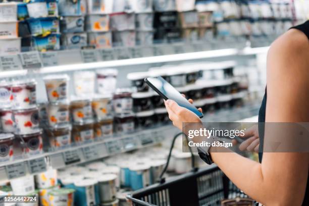 cropped shot of young asian woman holding shopping basket and checking her shopping list on smartphone while grocery shopping in a supermarket - shopping smartphone photos et images de collection