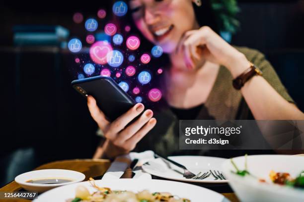 smiling young asian woman using smartphone on social media network application while having meal in the restaurant, viewing or giving likes, love, comment, friends and pages. social media addiction concept - kommunikation stock-fotos und bilder