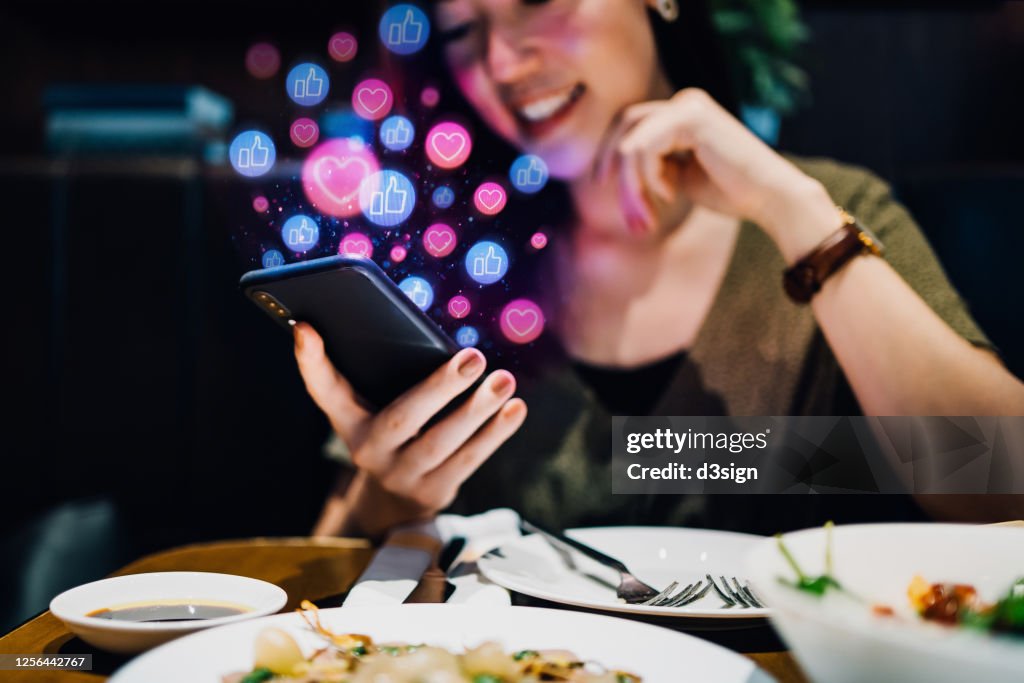 Smiling young Asian woman using smartphone on social media network application while having meal in the restaurant, viewing or giving likes, love, comment, friends and pages. Social media addiction concept