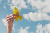 Hands holding yellow gold ribbon over blue sky, Sarcoma Awareness, Bone cancer, childhood cancer awareness, September yellow, World Suicide Prevention Day concept