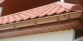 A close-up on red metal tile rooftop with wood soffit, house eaves, rafters and installed rain gutter.