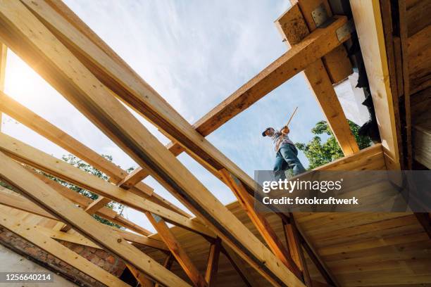 low angle view of mature male carpenter holding tape measure while standing on wooden roof beam at construction site against sky - building restoration stock pictures, royalty-free photos & images