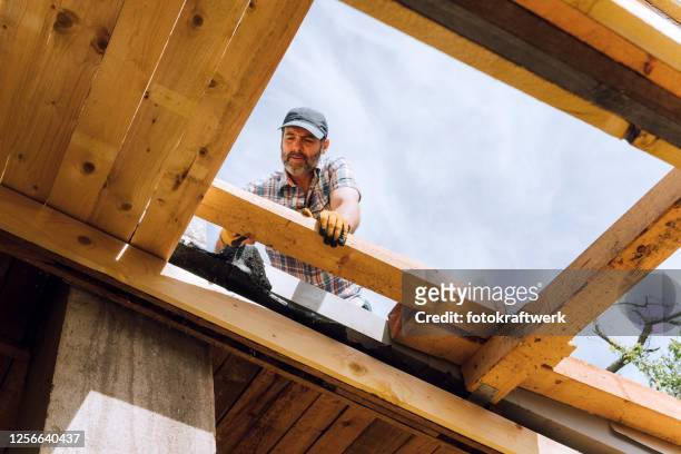 männlicher zimmermann kauert, während mit hammer auf holzdachbalken der unvollständigen wohnstruktur vor ort - bauen holz stock-fotos und bilder