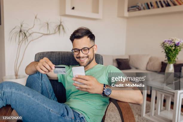 glücklicher junger mann beim online-shopping zu hause - credit card stock-fotos und bilder