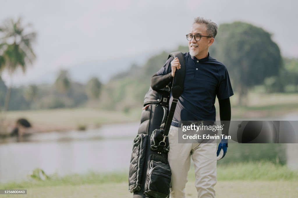 A matured chinese man golfer carrying a golf bag looking away in the golf course