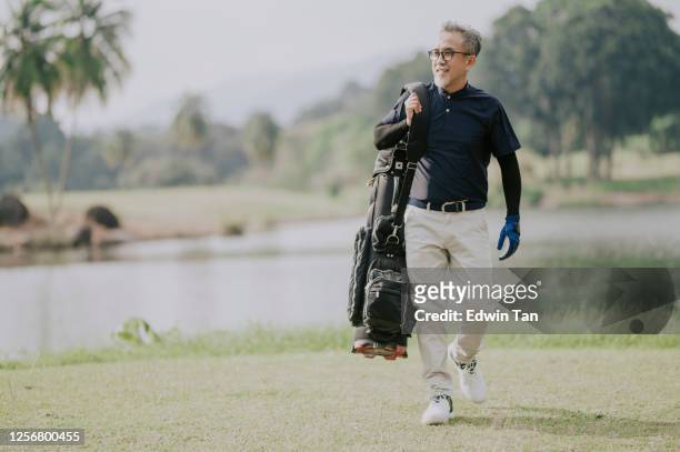 a matured chinese man golfer carrying a golf bag looking away in the golf course - golf bag stock pictures, royalty-free photos & images