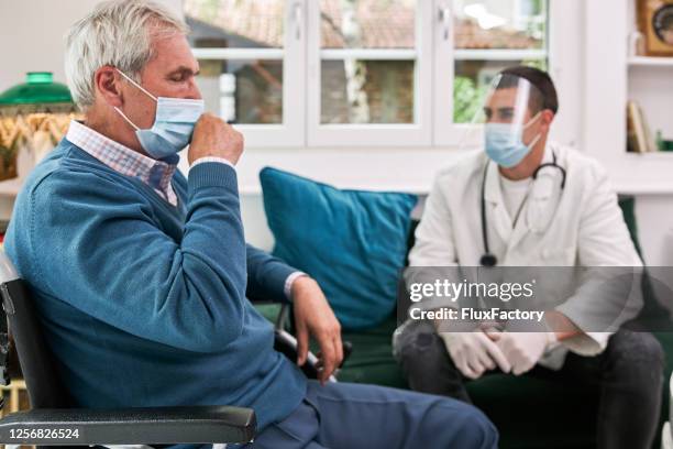 senior man coughing in a wheelchair while having a visit from a doctor - old cough stock pictures, royalty-free photos & images