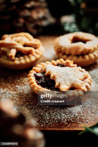 festive scene of 3 home made mince pies dusted with icing sugar on a rustic wooden board with pine cones and holly. - christmas mince pies stock pictures, royalty-free photos & images